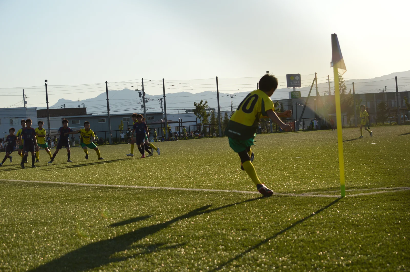 夕陽の差すピッチで、コーナーキックを蹴る真栄FCの選手